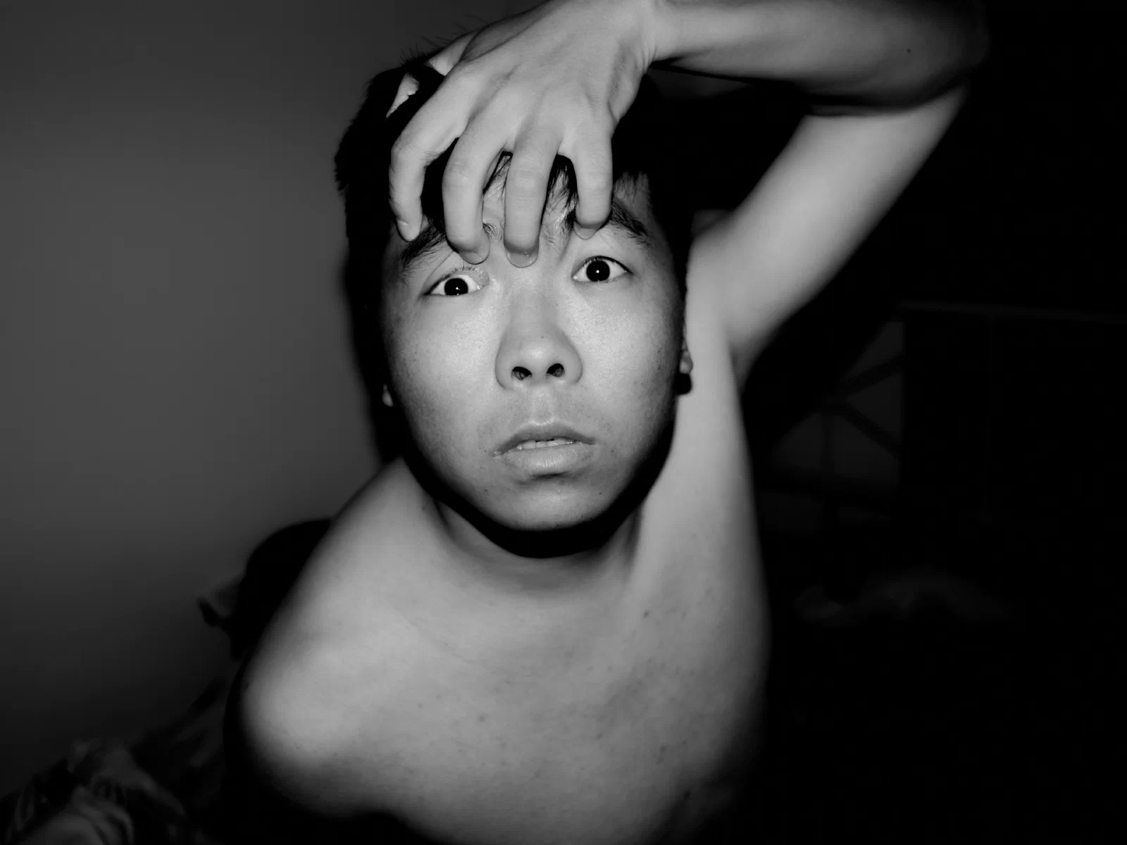 A bare-shouldered person looks directly into the camera with a hand pressed to the forehead.