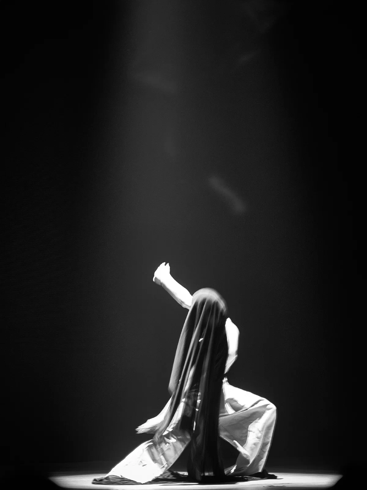 A kneeling performer raises an arm under a spotlight, the face hidden by black hanging fabric.