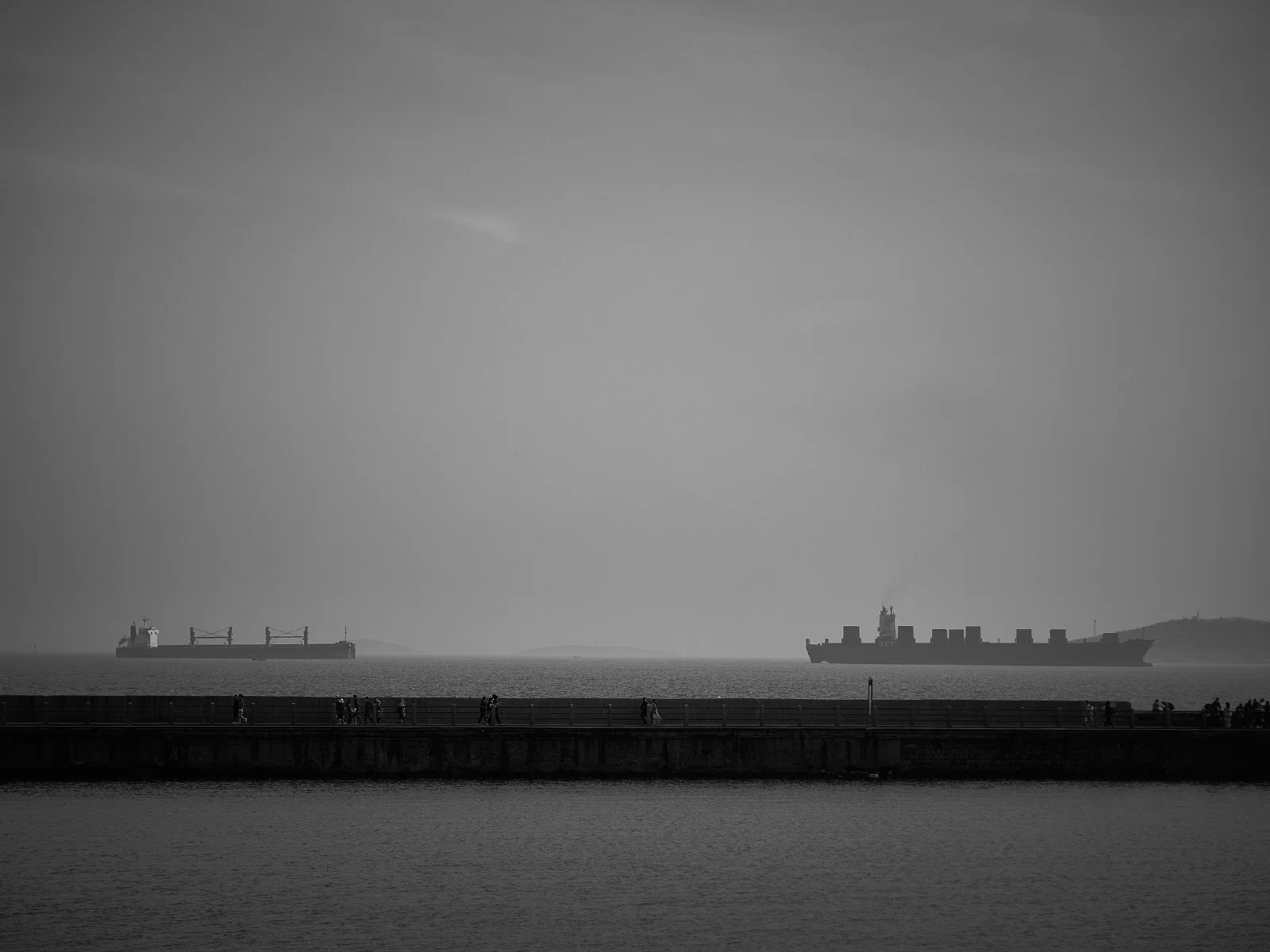 People walking along a seawall beneath distant cargo ships.