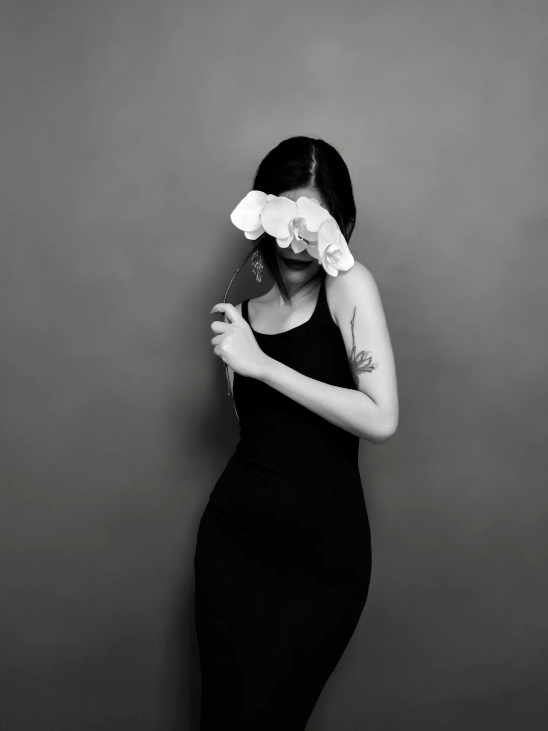 A person in a dark sleeveless dress holds a flowering stem, its pale blossoms covering the eyes.