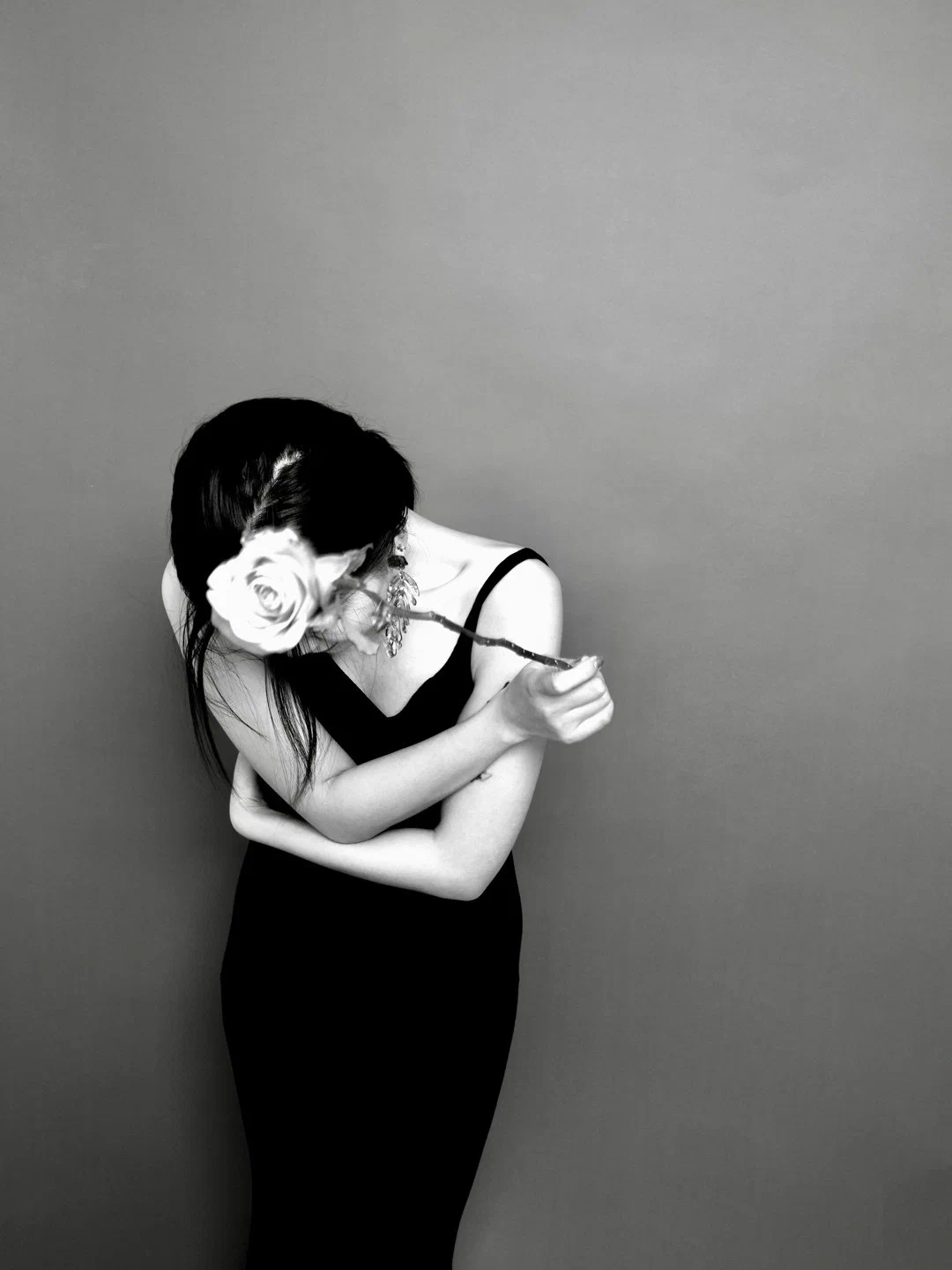 A person bows their head and crosses their arms, holding a rose in front of the face.